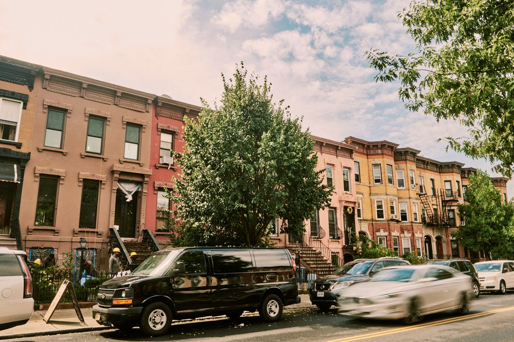 Parked cars along Halsey Street in Brooklyn. A white car drives by a parked van as construction workers renovate a brownstone.
