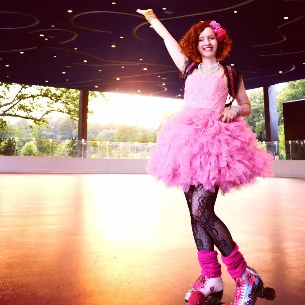 A woman in a pink tutu waves her arm in a rollerskating rink.