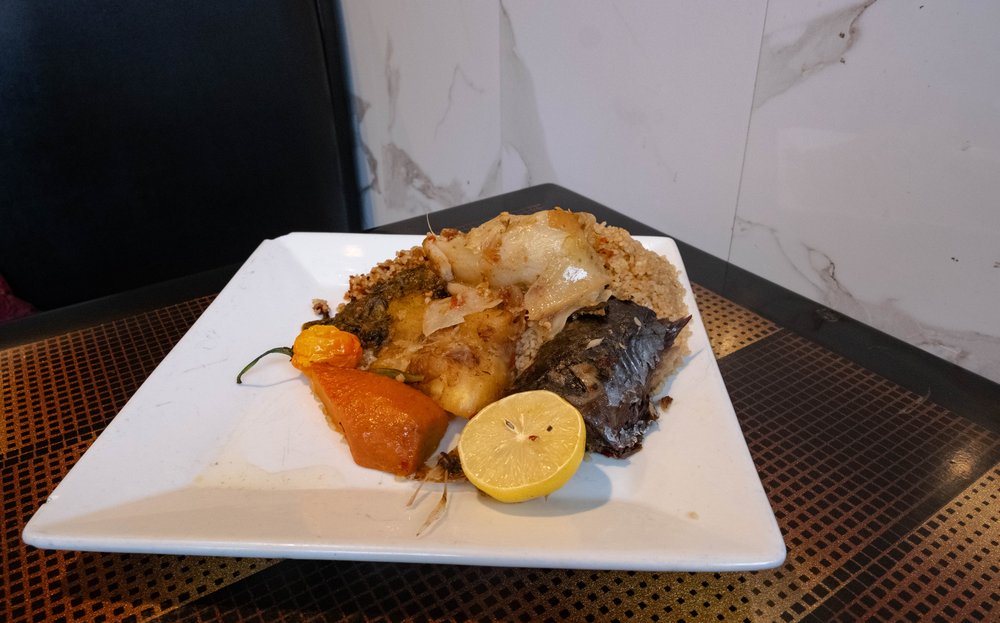 A plate of thiéboudienn is seen on a table. The dish contains jollof rice dish with whole vegetables, such as potatoes, cassava, and pumpkin, and flaky white fish.