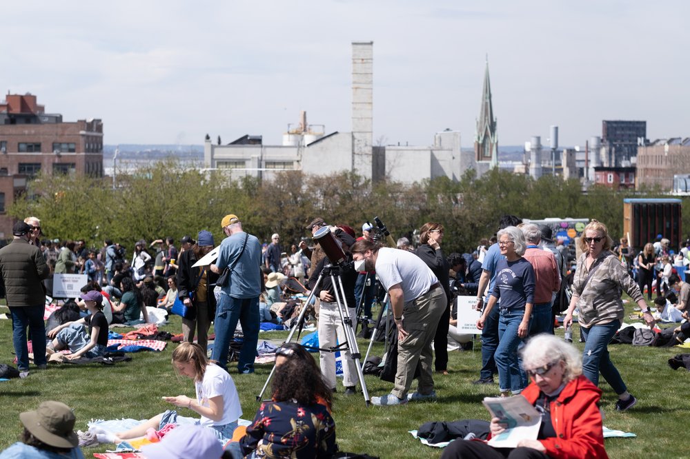 A view of a crowd at Green-Wood Cemetery in Brooklyn preparing to view the solar eclipse, with a scene of the New York City skyline in Brooklyn.