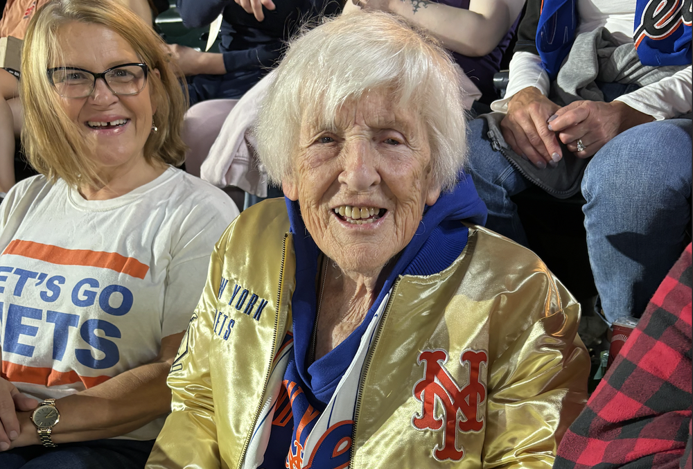 A woman smiles from the stands of a baseball game, wearing a Mets jacket.