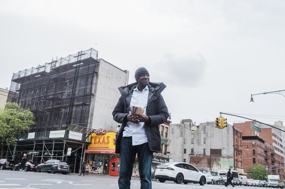 A man stands in the middle of the street, looking down the road, holding pamphlets.