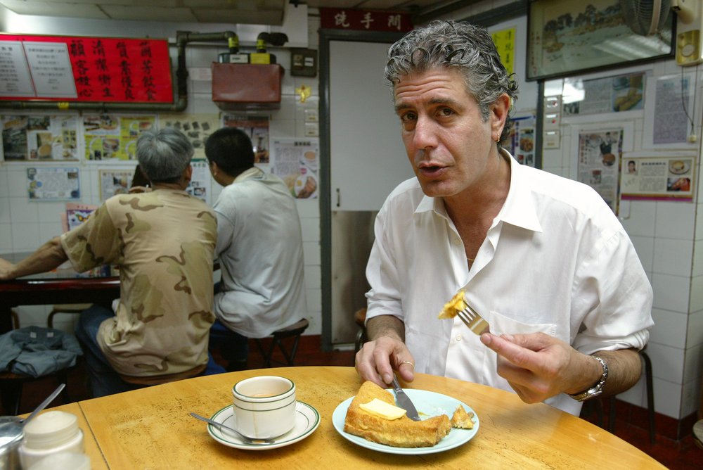 Anthony Bourdain sitting at a table, mid bite, in a restaurant in Hong Kong.