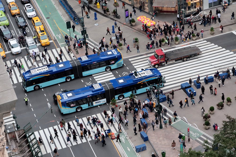 A bird's-eye view of two New York City buses at a busy intersection filled with pedestrians.