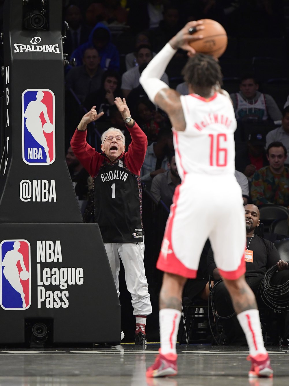 A basketball fan, seated behind the basket attempts to distract a player who is shooting a free throw.