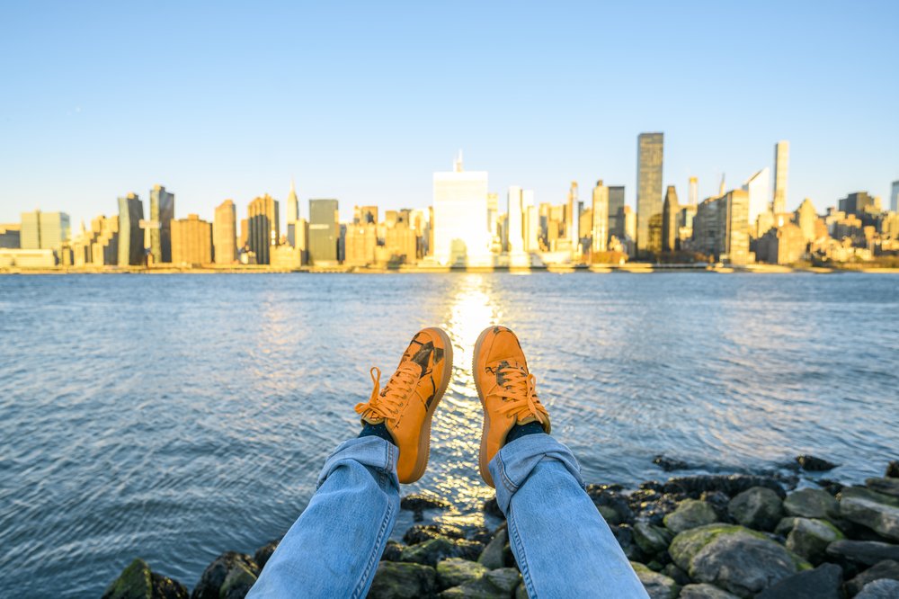 A person rests with their feet in orange sneakers, dangling over the water and looking at the NYC skyline.