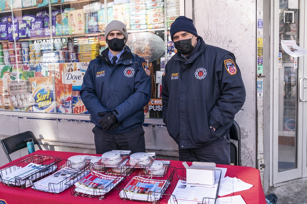 Two uniformed firefighters wearing face masks stand behind a table with fliers and smoke alarms on it.