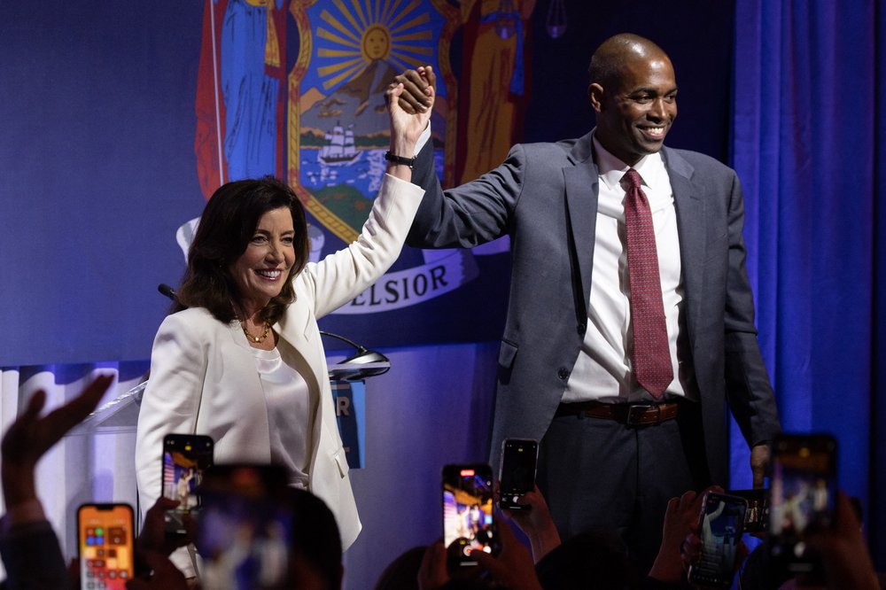 New York Governor Kathy Hochul and Lieutenant Governor Antonio Delgado stand holding hands before a flag that reads EXCELSIOR