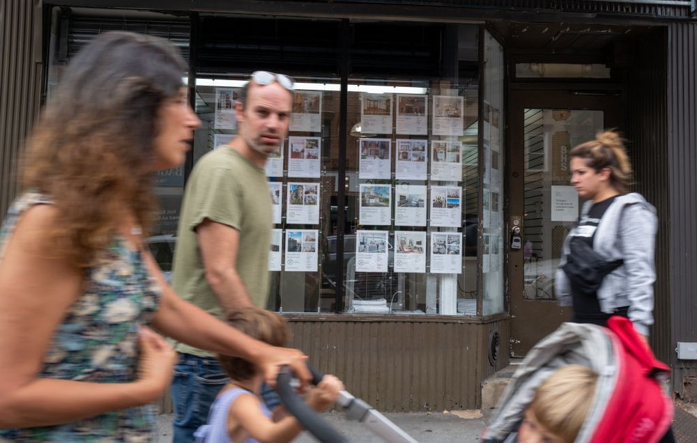 Rental listings are displayed on a realtor's office window as people walk by.