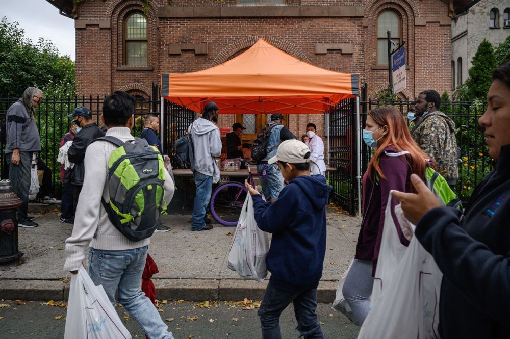 About a dozen people queue up along the side walk to receive food from a food pantry.