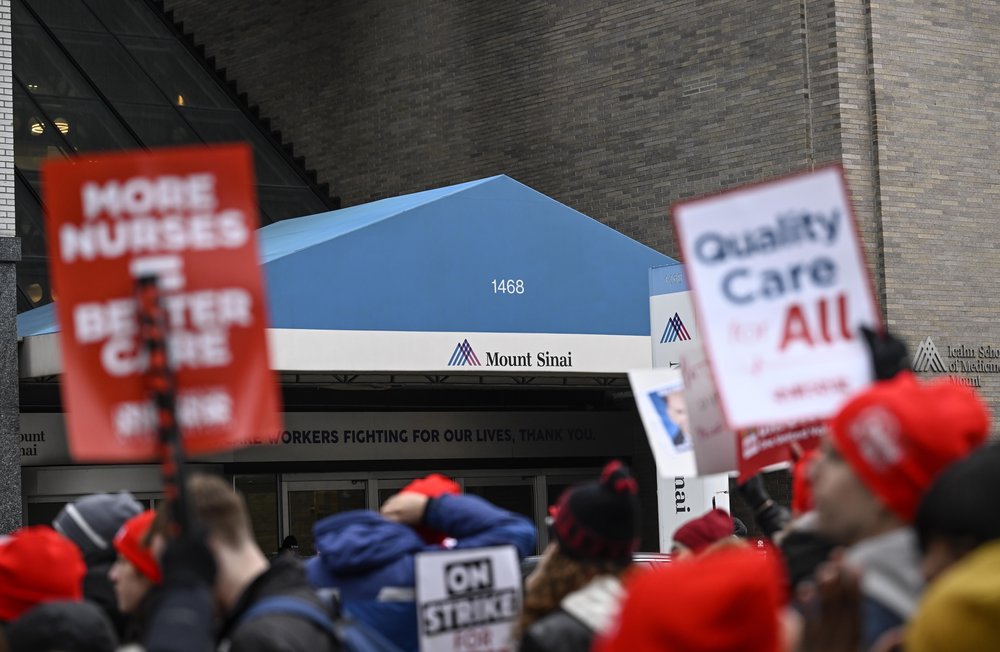 An entrance to Mt. Sinai Hospital with protesters out front holding signs, including one reading "quality care for all."