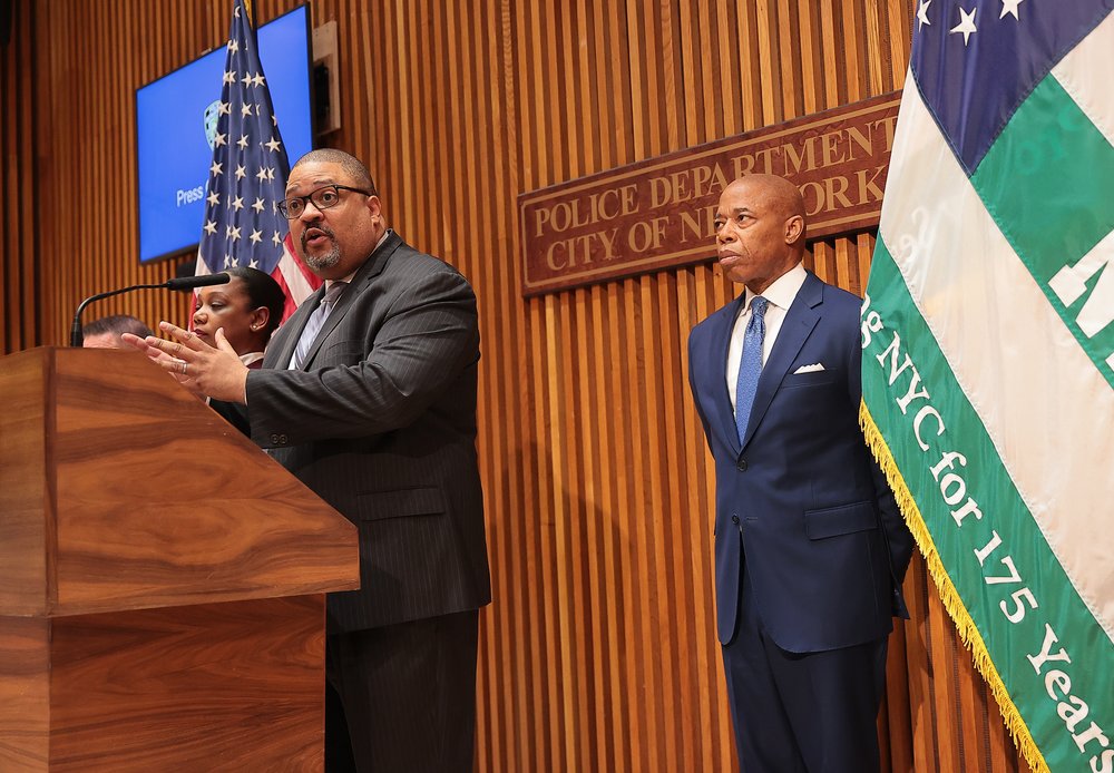 Manhattan District Attorney Alvin Bragg stands behind a wooden podium while Mayor Eric Adams stands in the background.