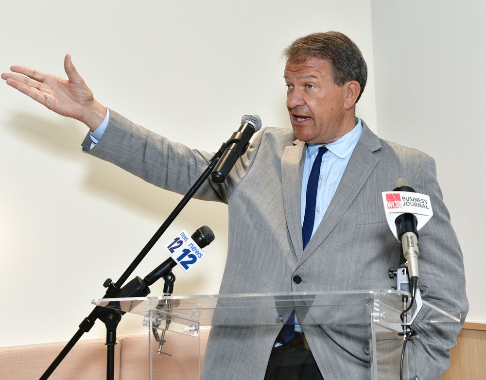 Westchester County Executive George Latimer holds up his hand as he stands behind a podium that has microphones on it.