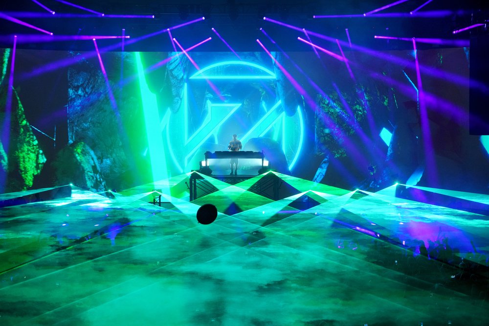 A DJ on stage with blue and green lights and purple lasers.