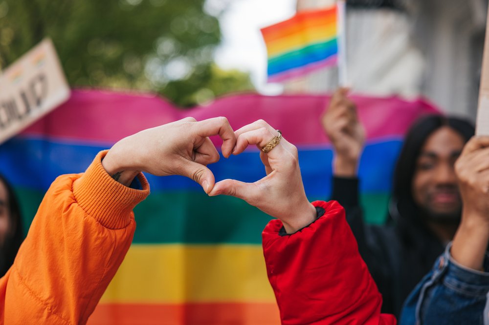 A person makes a heart with two hands in front of a rainbow flag.