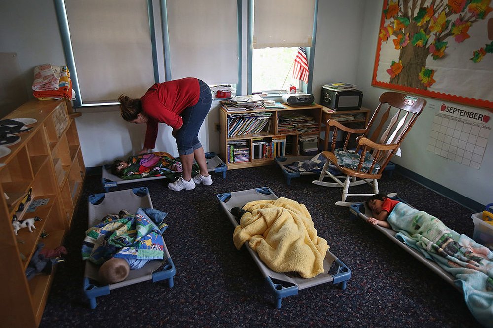 Children asleep on cots near the ground as a caretaker looks over them.