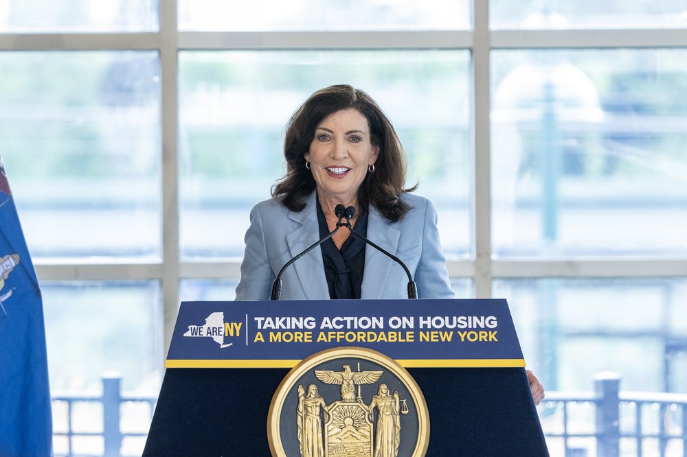 Gov. Kathy Hochul stands behind a podium with a sign that says "taking action on housing. A more affordable New York."