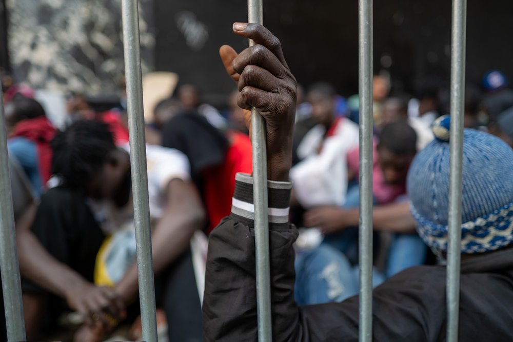 Dozens of recently arrived migrants to New York City camp outside of the Roosevelt Hotel on Aug. 1, 2023. A man, turned away from the camera, grabs a vertical rail in the foreground while looking toward a crowd of the gathered migrants.