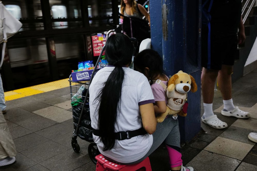 A young woman sits on a stool on the subway platform, holding her child as the pair sell candy on the subway.