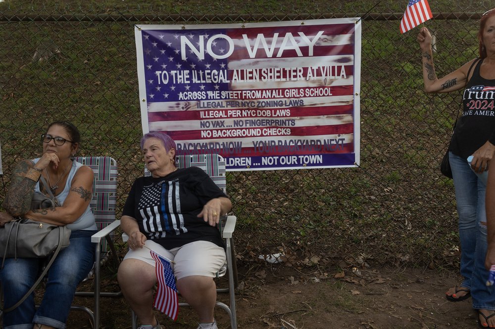 Two women sit in lawn chairs in front of a sign with an American flag and the message "No Way To the Illegal Alien Shelter."