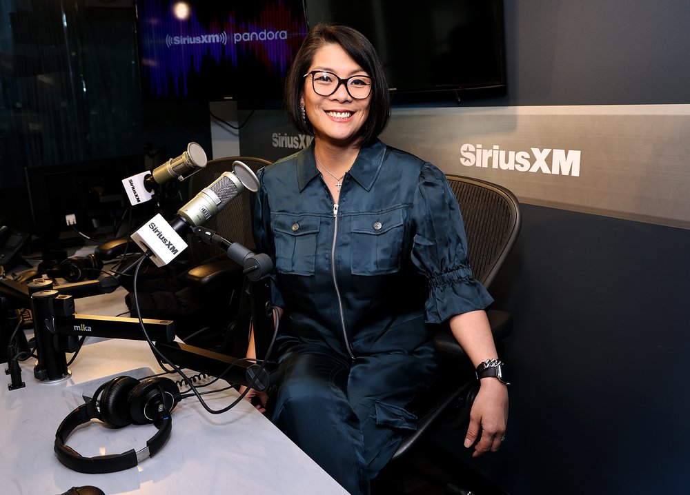 A woman wearing glasses and smiling while sitting at a microphone.