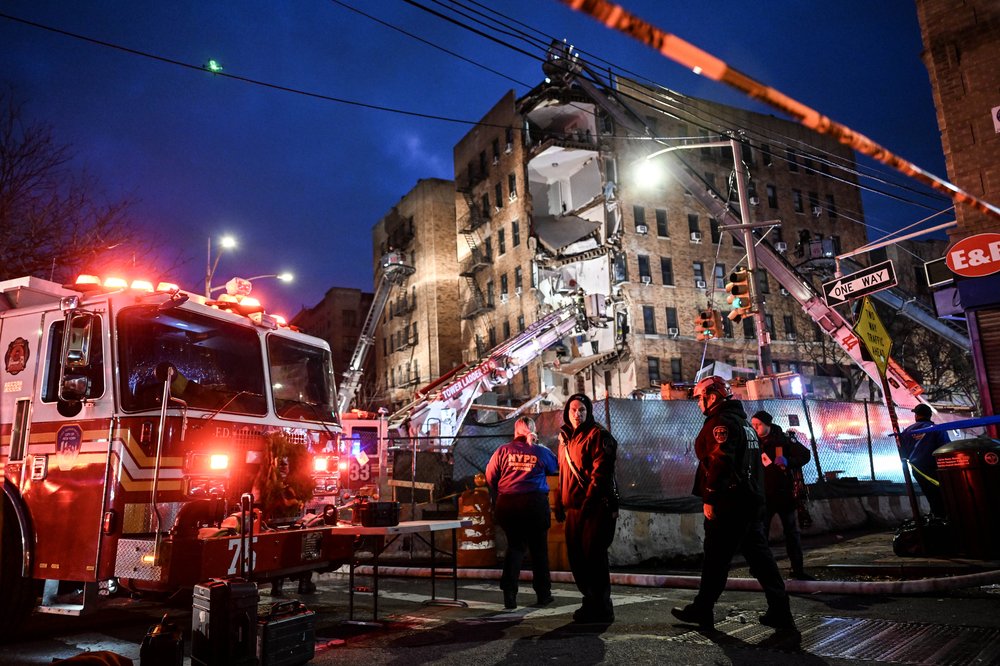 Firefighters and a fire engine are stationed on the street where a partial building collapse exposes the interior of a residential building.