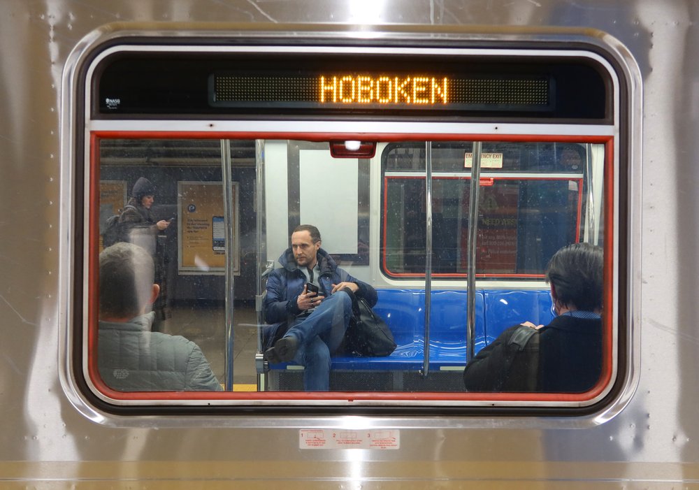 A train rider seen through a window, under a lighted sign that reads "Hoboken."