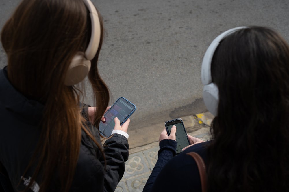 Two kids look at their smartphones while wearing headphones.