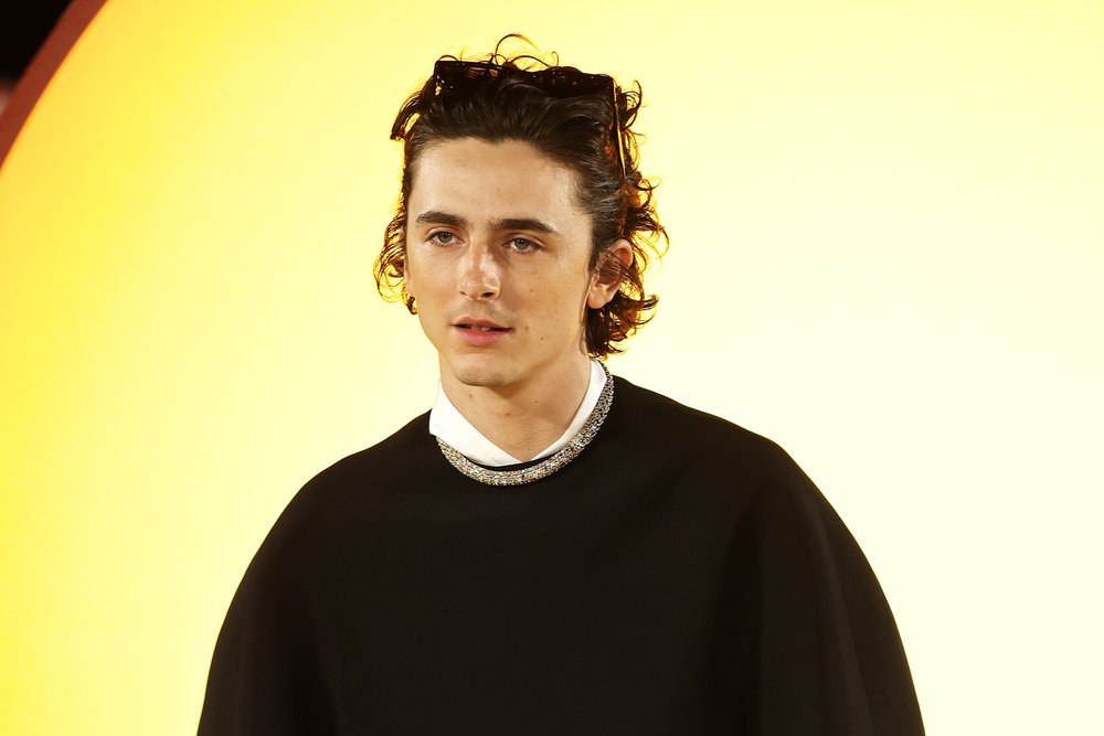 A man in a black sweater stands against a yellow background.