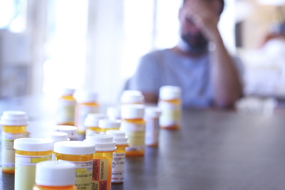 Pill bottles laid out on a table in front of a man holding his head.