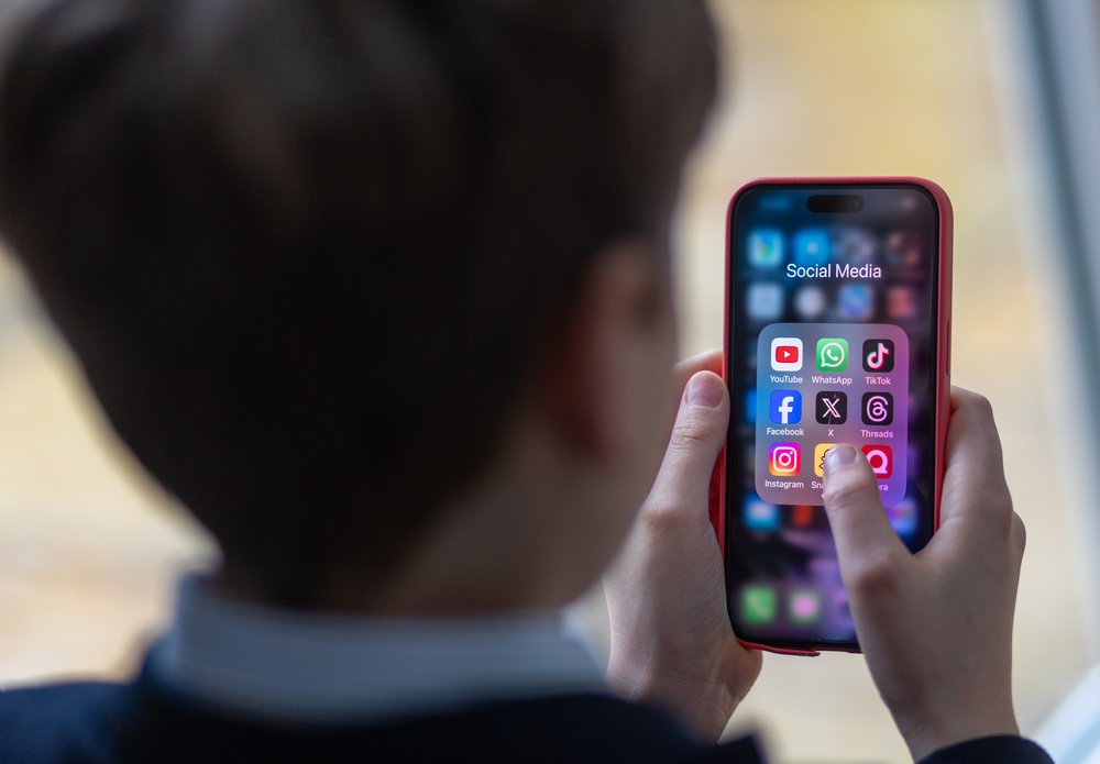 A young boy looks at a smartphone folder titled "Social Media" with apps such as TikTok and YouTube.