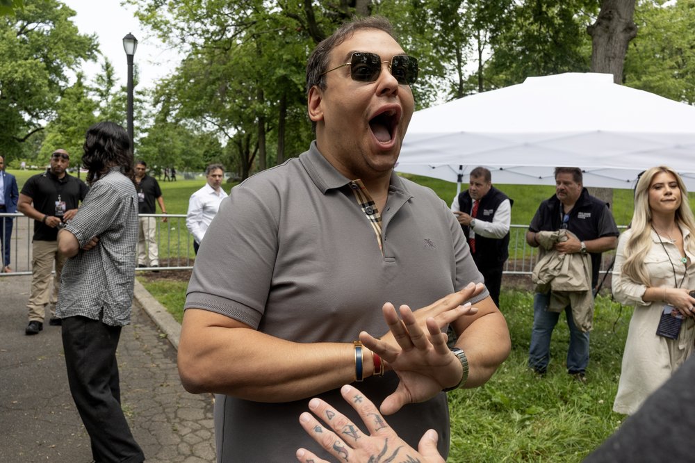 Former NY Rep. George Santos stands in sunglasses with his hands clapped against each other and his mouth open wide.