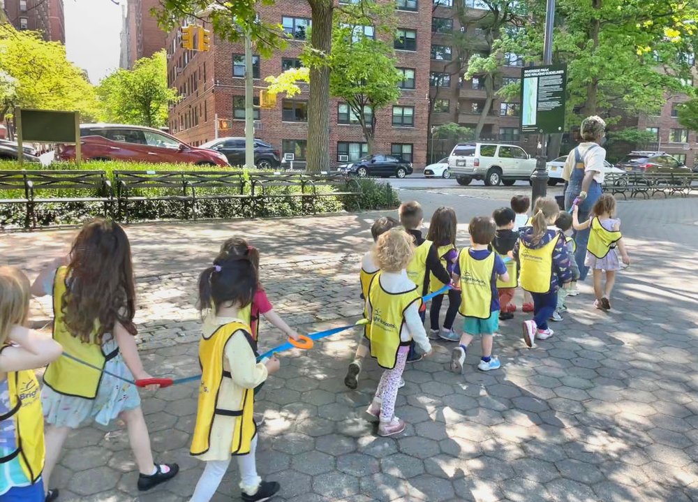 Children hold onto cords and wear yellow vests while they walk through a neighborhood with their daycare provider.