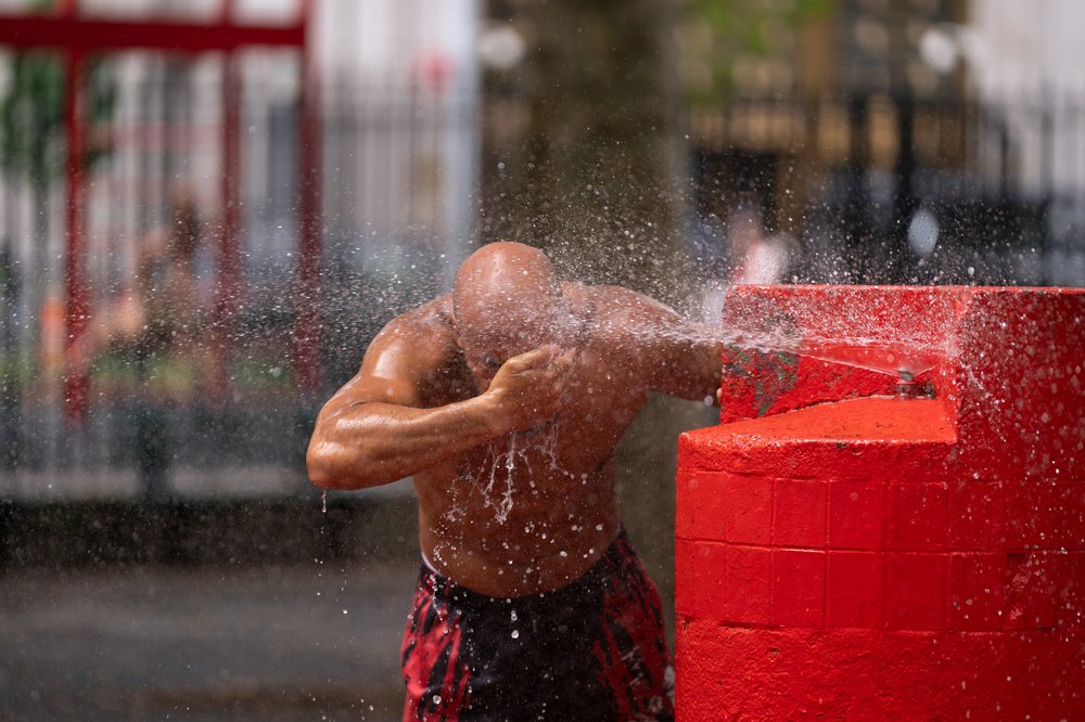 A shirtless man cools off with water spraying from a water fountain outdoors.