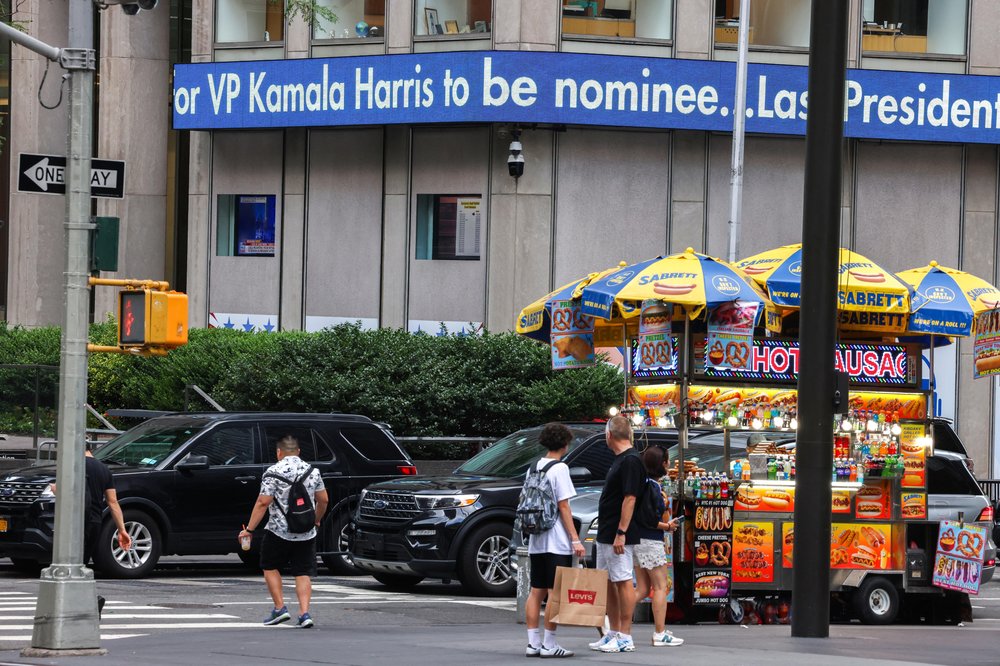 A ticker in New York City reads "VP Kamala Harris to be nominee" over a hot dog cart.