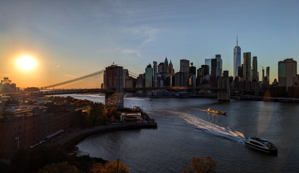 An NYC Ferry sails in the East River in front of the Brooklyn Bridge and lower Manhattan at sunset.