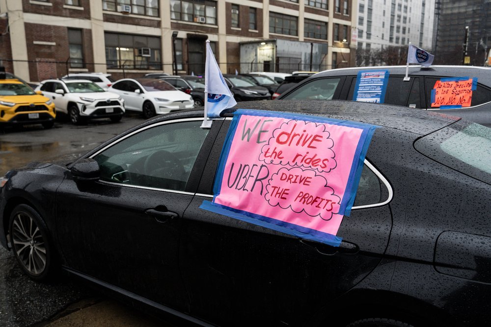 A black car with a pink signed pasted to its window that reads "We drive the rides, Uber drives the profits."