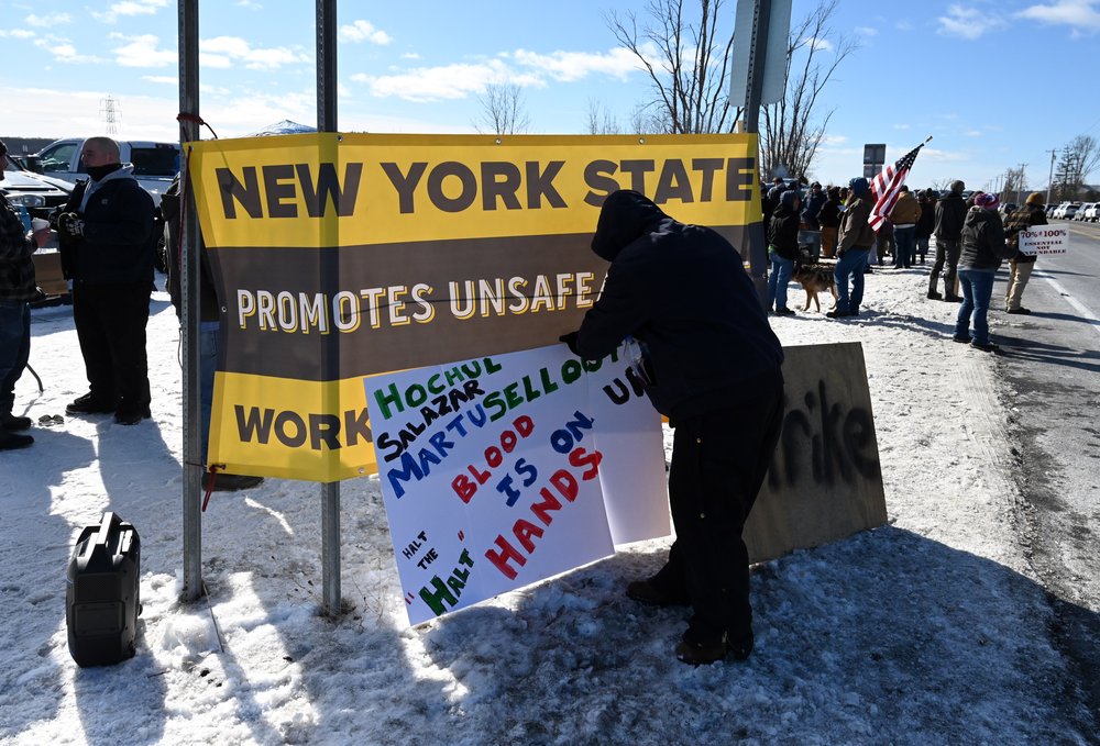 A man holds a sign that reads HOCHUL SALAZAR MARTUSELLOUT HALT THE "HALT"