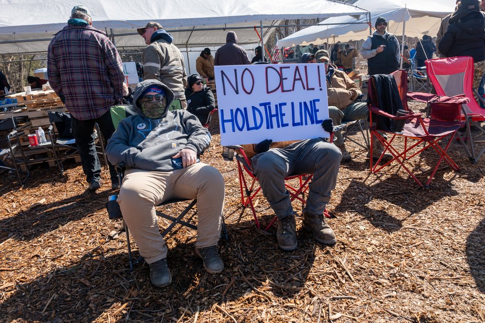 Men in folding chairs sit holding signs reading "NO DEAL HOLD THE LINE."
