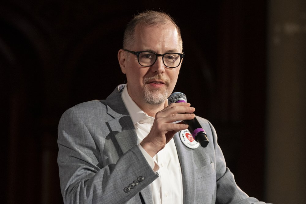 Mark Levine holds a microphone while wearing a grey suit and speaking at The Center at West Park.