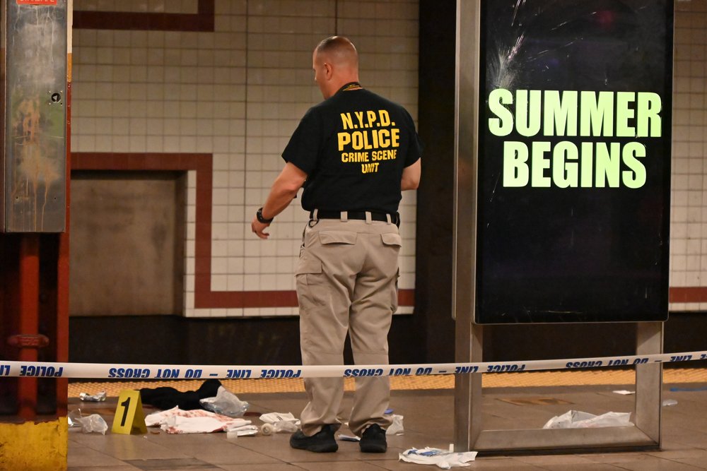 A police officer investigates a crime scene in the City Hall subway station next to a digital billboard that reads "Summer Begins."
