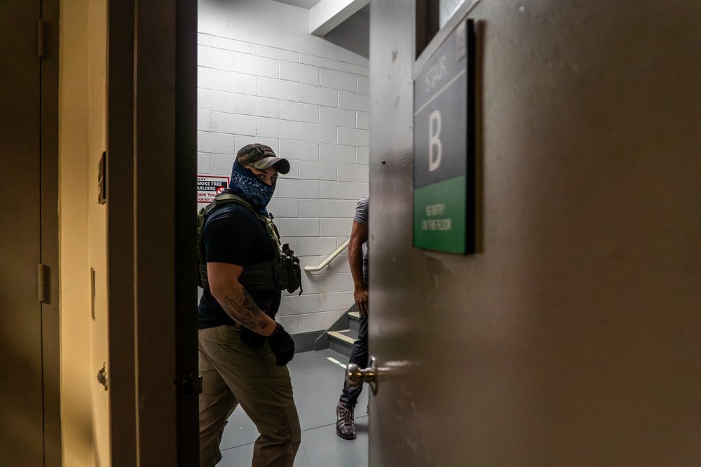 A masked federal agent looks directly at the camera as someone is detained and brought downstairs at 26 Federal Plaza.