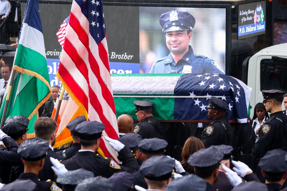 NYPD officers carry a casket with an American flag draped over it. A photo of Didarul Islam is seen on the screen in the background.