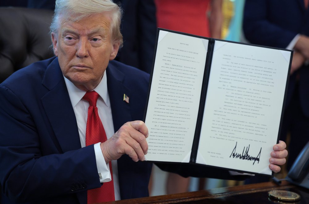 President Donald Trump sits at a desk holding up a signed executive order targeting cashless bail policies, with his bold signature visible at the bottom of the document.