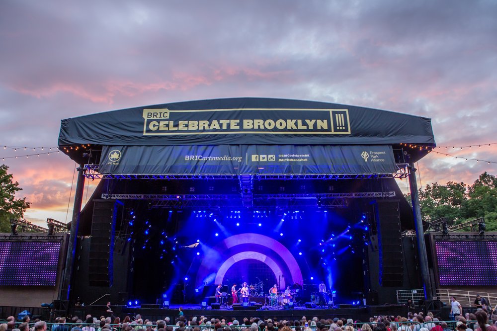 A large crowd of people at an outdoor concert with a black tarp over the stage, and a banner that reads "BRIC Celebrate Brooklyn!!!"