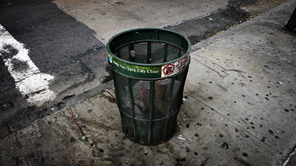 A dirty, old green mesh metal trash can on a New York City street.