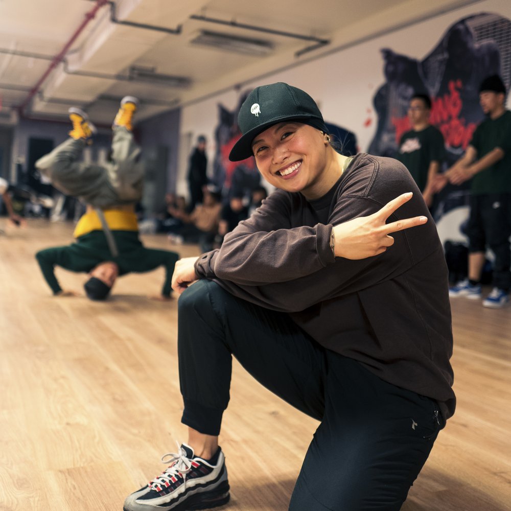 A dancer smiles for a photo, while sitting on one knee. Another dancer performs a headstand in the background.