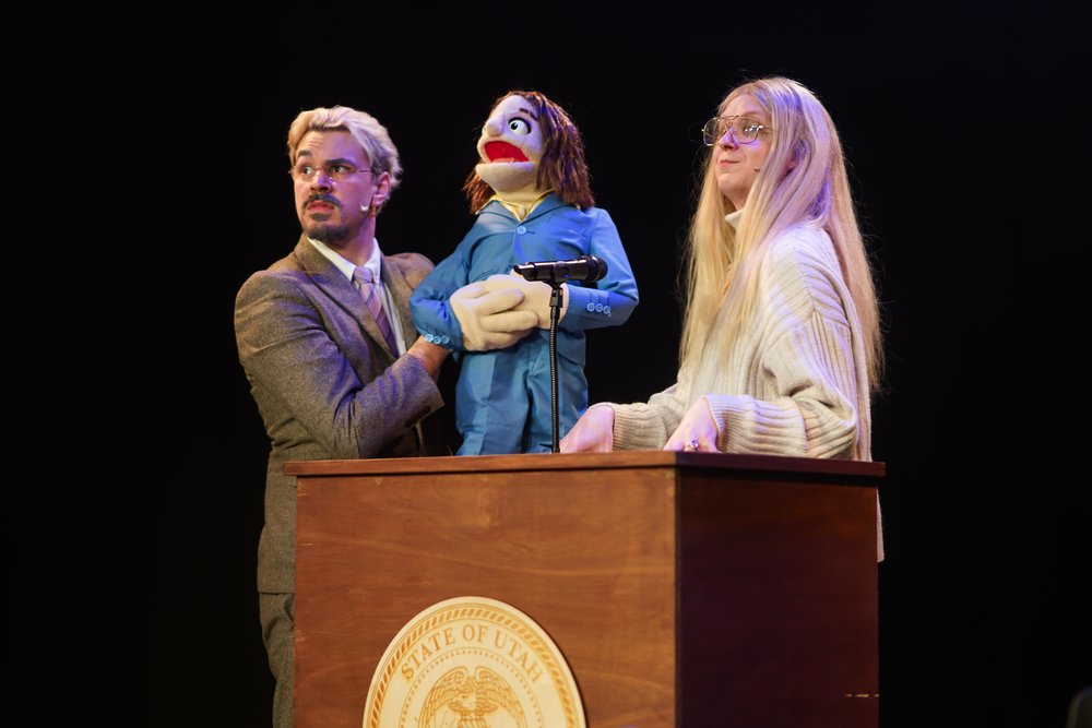 Two people stand behind a podium while one person holds a puppet.