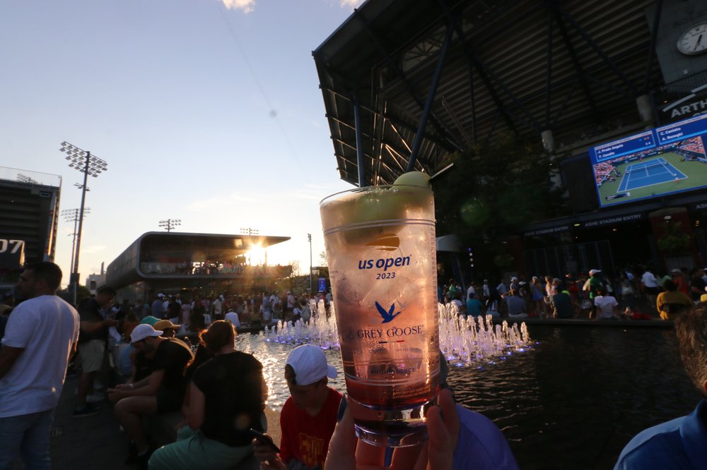 A photo of the pink Honey Deuce cocktail  with a water fountain and TV images of the U.S. Open in the background.