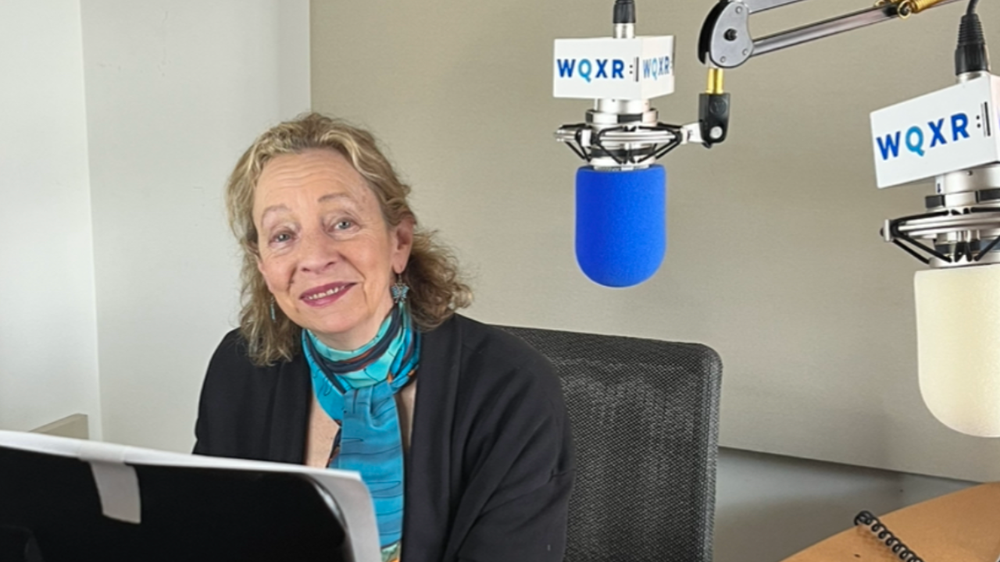 A woman sits at a chair with a microphone near her face.